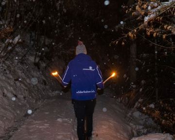 Eine Person läuft mit Fackeln auf einem verschneiten Weg in der Nacht. Es fällt Schnee und die Umgebung ist dunkel und ruhig.