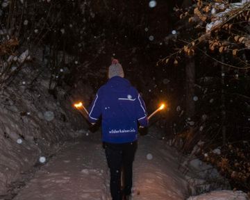 Eine Person läuft mit Fackeln auf einem verschneiten Weg in der Nacht. Es fällt Schnee und die Umgebung ist dunkel und ruhig.