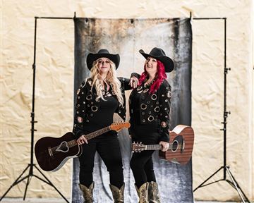 Zwei Musikerinnen stehen vor einem Hintergrund und halten Gitarren. Sie tragen schwarze Outfits und Hüte.