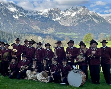 A brass band in traditional costumes stands in front of an impressive mountain backdrop. The musicians hold various instruments in their hands and smile at the camera.