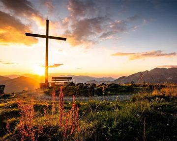 Ein wunderschöner Sonnenuntergang hinter einem Kreuz auf einem Berggipfel. Blumen und eine Bank im Vordergrund ergänzen die friedliche Landschaft.
