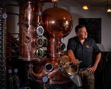 A man stands next to a large copper distillery in a distillery. The machine has various pressure gauges and a shiny surface.