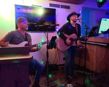 Ein Musiker spielt Gitarre und singt live, während ein anderer Musiker am Bass begleitet. Die Umgebung ist mit buntem Licht beleuchtet.