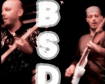 Two musicians are playing guitar on a stage. The background is dark and the letters "B S D" are prominently placed.