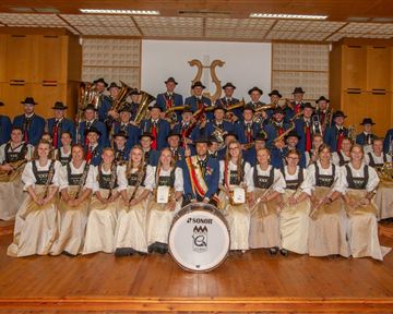 A group of musicians stands on a stage in traditional attire. They are surrounded by musical instruments and proudly present their ensemble.