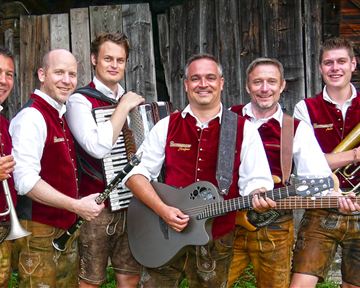 Sechs Musiker in traditionellen Trachten stehen vor einer Holzwand. Sie halten verschiedene Instrumente und lächeln in die Kamera.