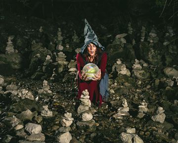 Eine mystische Frau trägt einen Zauberhut und hält eine glühende Kugel. Um sie herum sind viele steinerne Türmchen, die eine geheimnisvolle Atmosphäre schaffen.