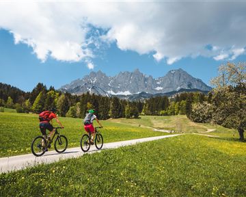 Zwei Radfahrer fahren auf einem Weg durch eine grüne Wiese. Im Hintergrund sind beeindruckende Berge und ein blauer Himmel zu sehen.