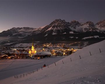 A picturesque alpine landscape at night with snow-covered mountains. In the foreground, a small village with illuminated houses and a church is visible.