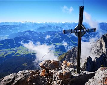 Ein beeindruckendes Bergpanorama mit einem Kreuz auf einem Gipfel. Im Hintergrund sind majestätische Berge und ein klarer Himmel zu sehen.