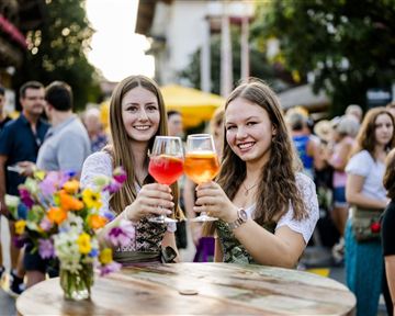 Two young women toast with colorful drinks. In the background, people and market stalls can be seen, creating a festive atmosphere.
