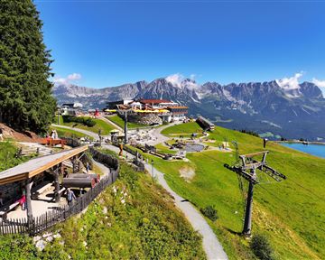 Eine malerische Berglandschaft mit einem Restaurant und Aussicht auf die umliegenden Berge. Im Vordergrund sind Wanderwege und eine Seilbahn zu sehen.