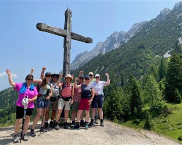 Eine Gruppe von Wanderern steht vor einem großen Holzkreuz in den Bergen. Sie lächeln und posieren fröhlich auf einem sonnigen Tag.