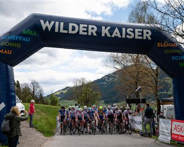 A bicycle race begins under a large arch with the inscription "Wilder Kaiser". In the background are mountain landscapes and the participants ready to start.