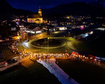 A festive event at night with many people carrying lights and lanterns. In the background, a church and a small town view can be seen.