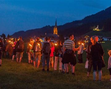 Eine Gruppe von Menschen mit Fackeln geht durch eine Wiese bei Dämmerung. Im Hintergrund ist eine Kirche und eine malerische Landschaft zu sehen.