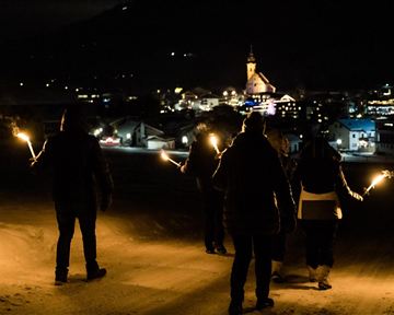 Eine Gruppe von Menschen steht nachts im Schnee und hält Fackeln in der Hand. Im Hintergrund ist eine beleuchtete Stadt mit einer Kirche zu sehen.
