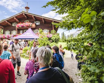 A group of people stands in front of a traditional wooden house with blooming windows. In the background, there is a green landscape that highlights the beautiful day.