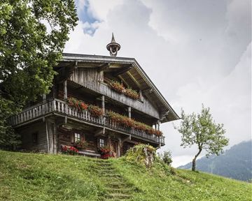 A traditional wooden house with a balcony and colorful flowers. It stands in a green landscape under a cloudy sky.