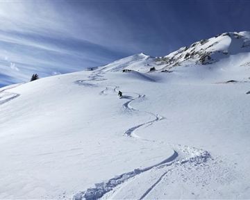 Eine schneebedeckte Berglandschaft mit einem Skifahrer, der eine Spur im frischen Schnee hinterlässt. Der Himmel ist klar und blau, was ein schönes Winterbild schafft.