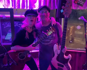 Two musicians stand together on a stage. They hold instruments and smile at the camera while the surroundings are illuminated with colorful lights.