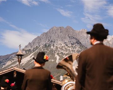 Two people in traditional clothing stand against a majestic mountain backdrop. The sky is clear and the landscape is characterized by a charming rural ambiance.