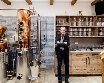 Eine moderne Brennerei mit Kupferkessel und Holzregalen. Ein Mann steht entspannt neben der Ausrüstung.