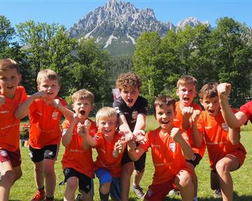 Eine Gruppe fröhlicher Kinder in orangefarbener Sportkleidung posiert auf einem grünen Feld. Im Hintergrund sind hohe Berge und ein blauer Himmel zu sehen.