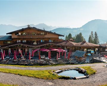 Ein malerisches Alpenhaus mit einer einladenden Terrasse und Blick auf die Berge. Die Umgebung ist grün und sonnig, ideal für einen entspannten Aufenthalt.