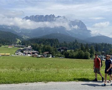 Two people are walking on a path in a green landscape. In the background, majestic mountains rise up with clouds.