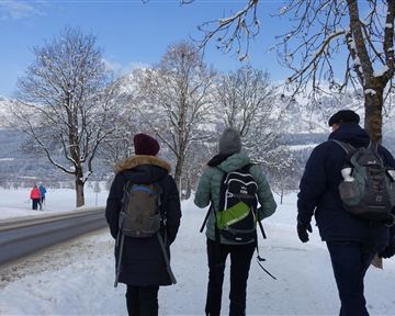 Drei Personen wandern auf einem schneebedeckten Weg. Im Hintergrund sind schneebedeckte Berge und Bäume zu sehen.