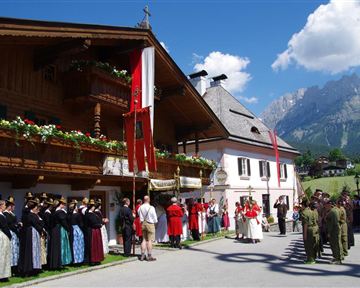 A traditional celebration in a picturesque village. People in festive attire stand together, surrounded by historic buildings and mountains.