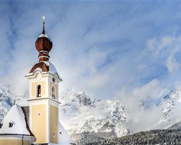 Eine hübsche Kirche mit einer charakteristischen Kuppel steht im Schnee. Im Hintergrund sind majestätische Berge und ein blauer Himmel zu sehen.