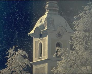 A snowy church tower with an elegant roof stands between snow-covered trees. In the background, a clear winter sky can be seen.