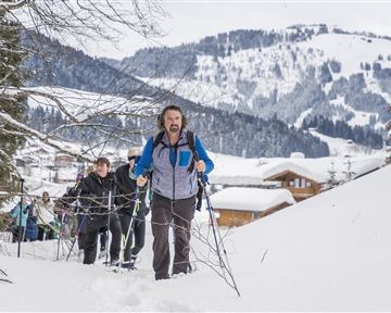 A group of hikers is climbing a snow-covered slope in the mountains. The surroundings are wintry with snow-covered trees and cabins in the background.