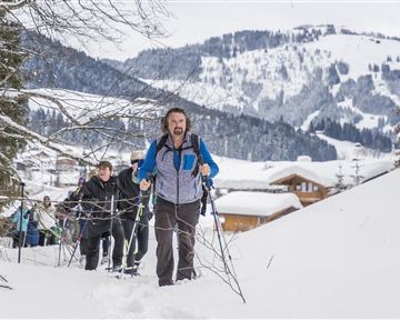 Eine Gruppe von Wanderern erklimmt einen schneebedeckten Hang in den Bergen. Die Umgebung ist winterlich mit schneebedeckten Bäumen und Hütten im Hintergrund.