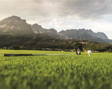 Ein Golfplatz mit grünem Gras und einem Golfball auf einem Tee. Im Hintergrund sind beeindruckende Berge und ein bewölkter Himmel zu sehen.