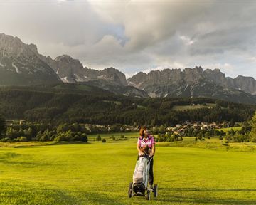 Eine Frau mit einem Golfwagen steht auf einem grünen Golfplatz. Im Hintergrund sind beeindruckende Berge und ein bewölkter Himmel zu sehen.