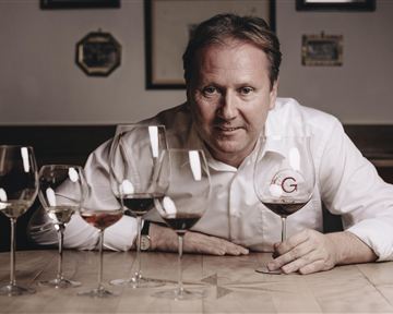 A man is sitting at a table and holding a wine glass in his hand. In front of him are five wine glasses with different types of wine.