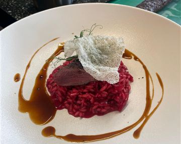 Ein kunstvoll angerichtetes Gericht mit rotem Risotto, zartem Fleisch und einer knusprigen Dekoration. Eine dekorative Sauce umrahmt das Ganze auf dem Teller.