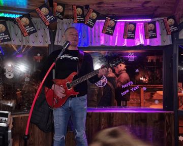 Ein Musiker mit einer roten Gitarre steht auf einer kleinen Bühne und singt. Im Hintergrund sind bunte Lichter und festliche Dekorationen zu sehen.