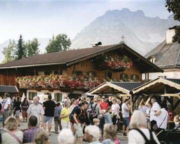 A bustling marketplace with many people in front of a traditional wooden house. In the background, mountains and trees can be seen, creating an idyllic atmosphere.