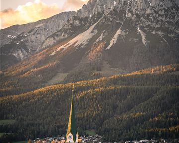 Eine malerische Landschaft mit grünen Wiesen und beeindruckenden Bergen im Hintergrund. Im Vordergrund ist ein kleiner Ort mit einer Kirche sichtbar.