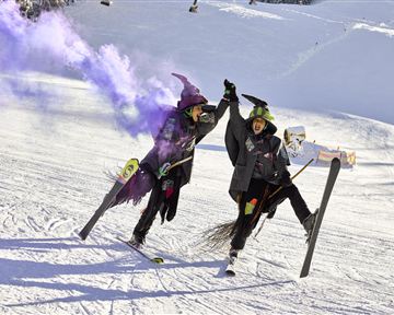Zwei verkleidete Hexen feiern auf der Skipiste und halten sich an den Händen. Bunte Rauchfackeln umgeben sie in der verschneiten Landschaft.