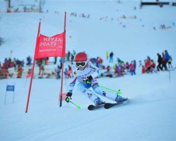 Ein Skifahrer fährt durch ein Tor auf einer verschneiten Piste. Im Hintergrund sind viele Zuschauer und weitere Skifahrer zu sehen.