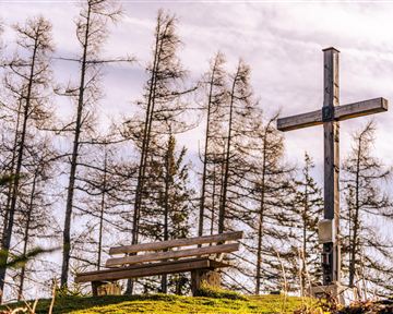 Eine Holzbank steht neben einem Holzkreuz, umgeben von kahlen Bäumen. Der Himmel ist leicht bewölkt.