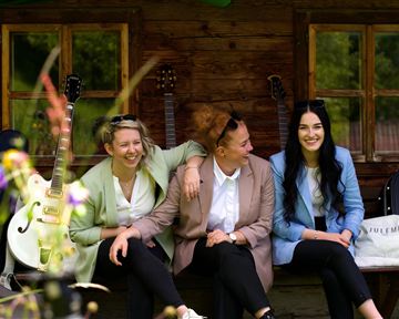 Three women are sitting happily on a bench in front of a wooden house. In the background, guitars and plants can be seen.