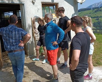 Eine Gruppe von Menschen steht draußen vor einem Bauernhof. Sie schauen sich neugierig etwas an und scheinen interessiert zu sein.