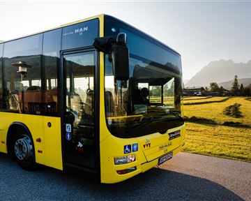 Ein gelber Bus steht auf einer Straße neben einem Feld. Im Hintergrund sind Berge und ein klarer Himmel zu sehen.