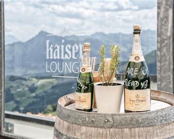 Eine rustikale Barrique-Fass mit zwei Champagnerflaschen und einer kleinen Pflanze. Im Hintergrund sind Berge und eine Fensterbeschriftung zu sehen.